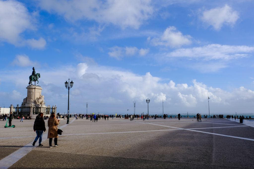 People walking across the open riverside square of Pra&ccedil;a do Com&eacute;rcio under a wide blue sky, Lisbon