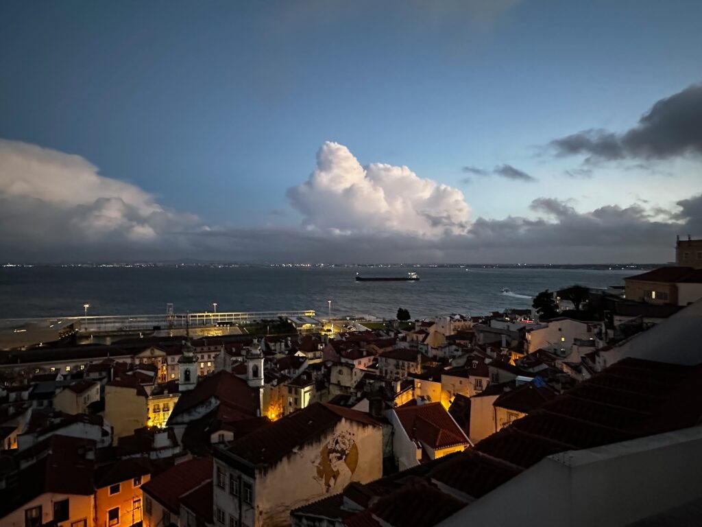 Lisbon rooftops and the Tagus River at dusk, city lights under a cloudy evening sky