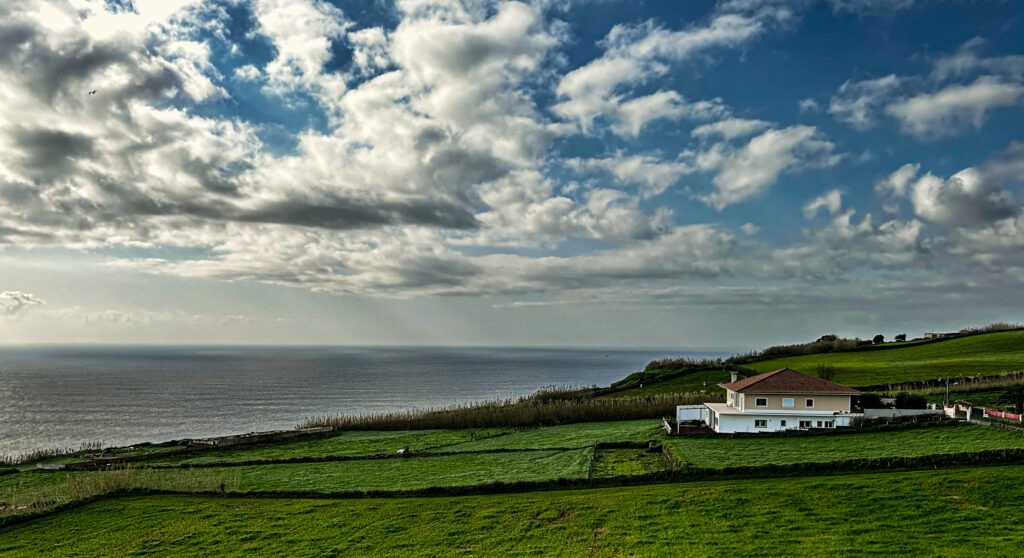 Green coastal farmland with a white farmhouse above the Atlantic under drifting clouds, Azores