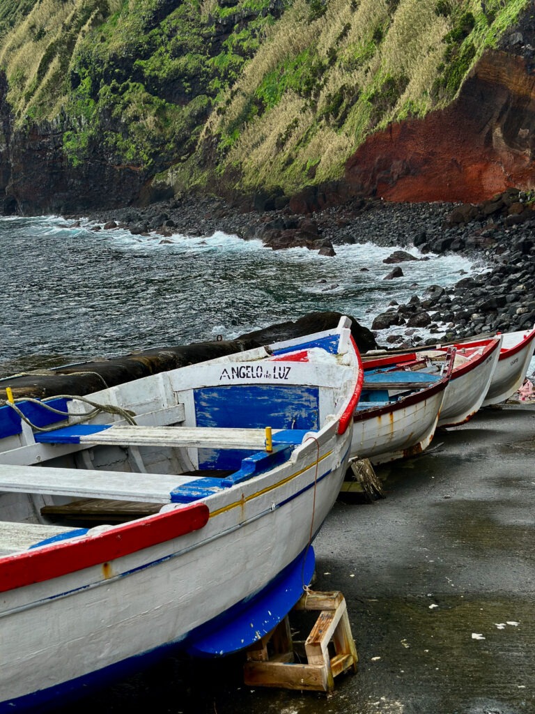 Painted wooden fishing boats on a rocky shore below green and red volcanic cliffs, Azores