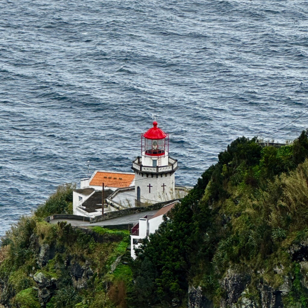 White lighthouse with a red lantern on a green cliff above the Atlantic, Azores