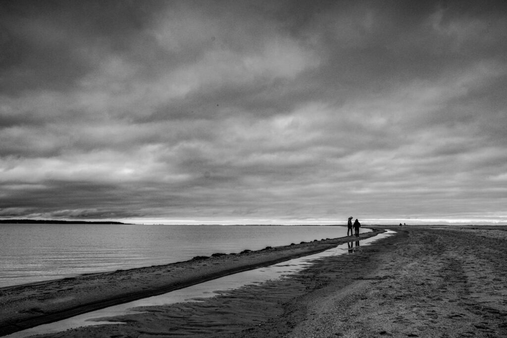 Long narrow sandbar stretching into the sea beneath heavy clouds, black and white minimalist seascape